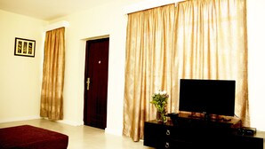 A room with a dark wooden door, a large window with golden curtains, a flat-screen TV on a black stand, and a red rug on the floor.