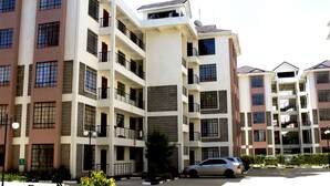 A multi-story residential building with balconies, a parking area, and a small tree.