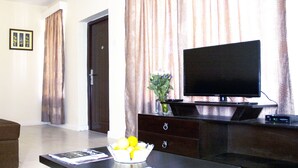 A living room with a flat-screen TV on a wooden stand, a coffee table with magazines, a brown sofa, and a door in the background.