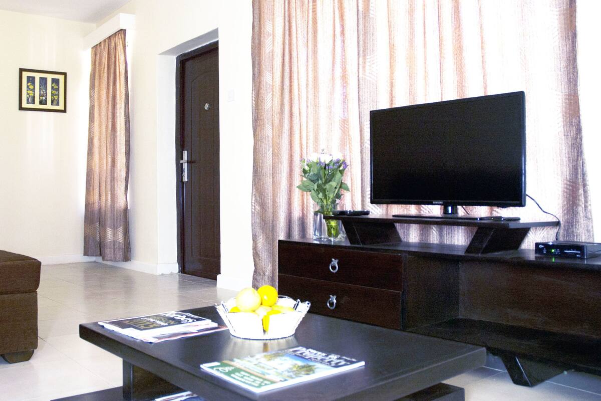 A living room with a flat-screen TV on a wooden stand, a coffee table with magazines, a brown sofa, and a door in the background.