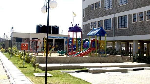 A playground area with slides and climbing structures, surrounded by a paved walkway and a building with multiple windows.