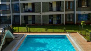 A swimming pool with a clear blue water surface, surrounded by a tiled deck. In the background, there is a multi-story building with balconies and windows.