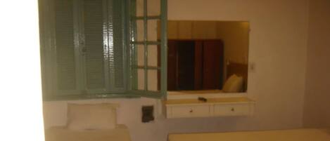 A room with two single beds, a small window with green shutters, a mirror above the bed, and a small dresser with a mirror.