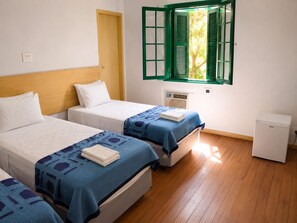A hotel room with two beds, a small bedside table, a window with green shutters, and a compact refrigerator.