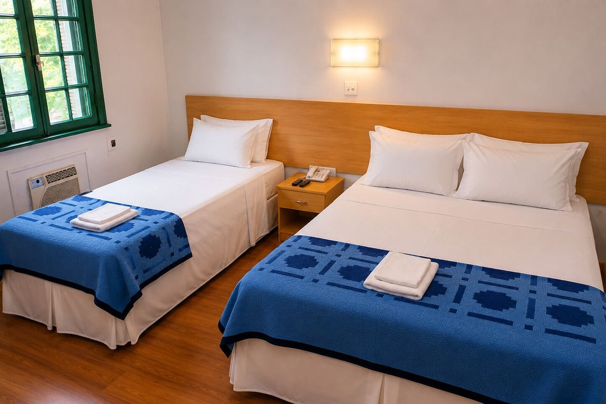 A hotel room with two beds, each with a blue and white bedspread, a wooden headboard, a bedside table with a phone, and a window with green frames.