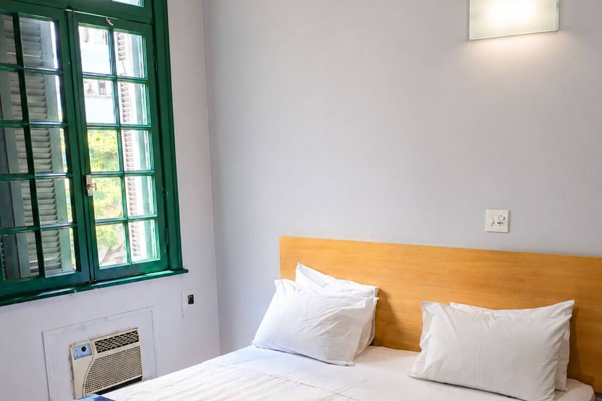 A hotel room with a bed, a window with green shutters, a wall-mounted light, and a radiator.