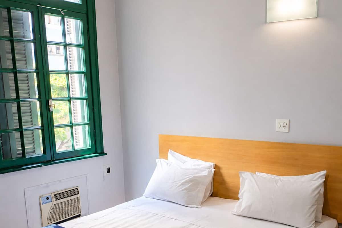 A hotel room with a bed, a window with green shutters, a wall-mounted light, and a radiator.