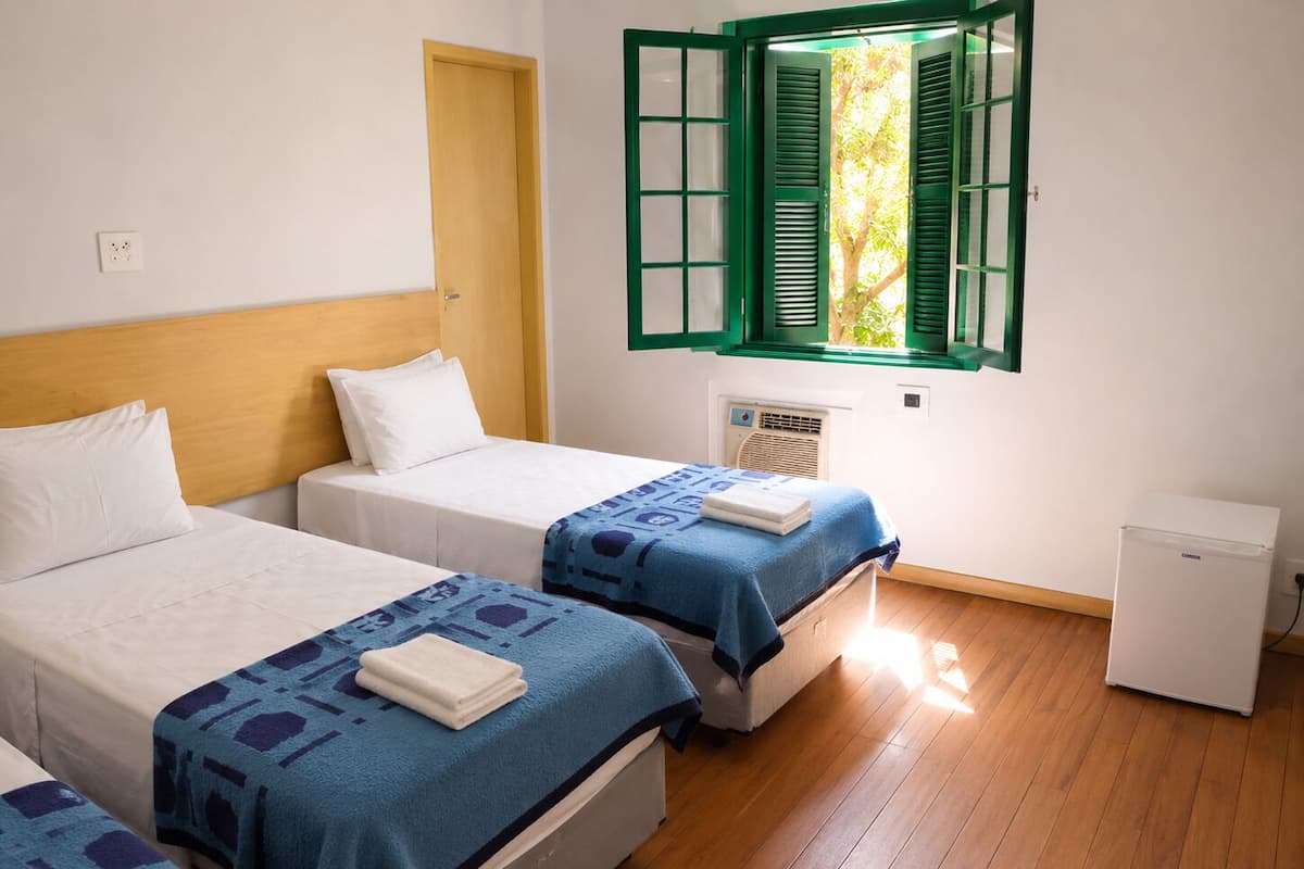 A hotel room with two beds, a small bedside table, a window with green shutters, and a compact refrigerator.