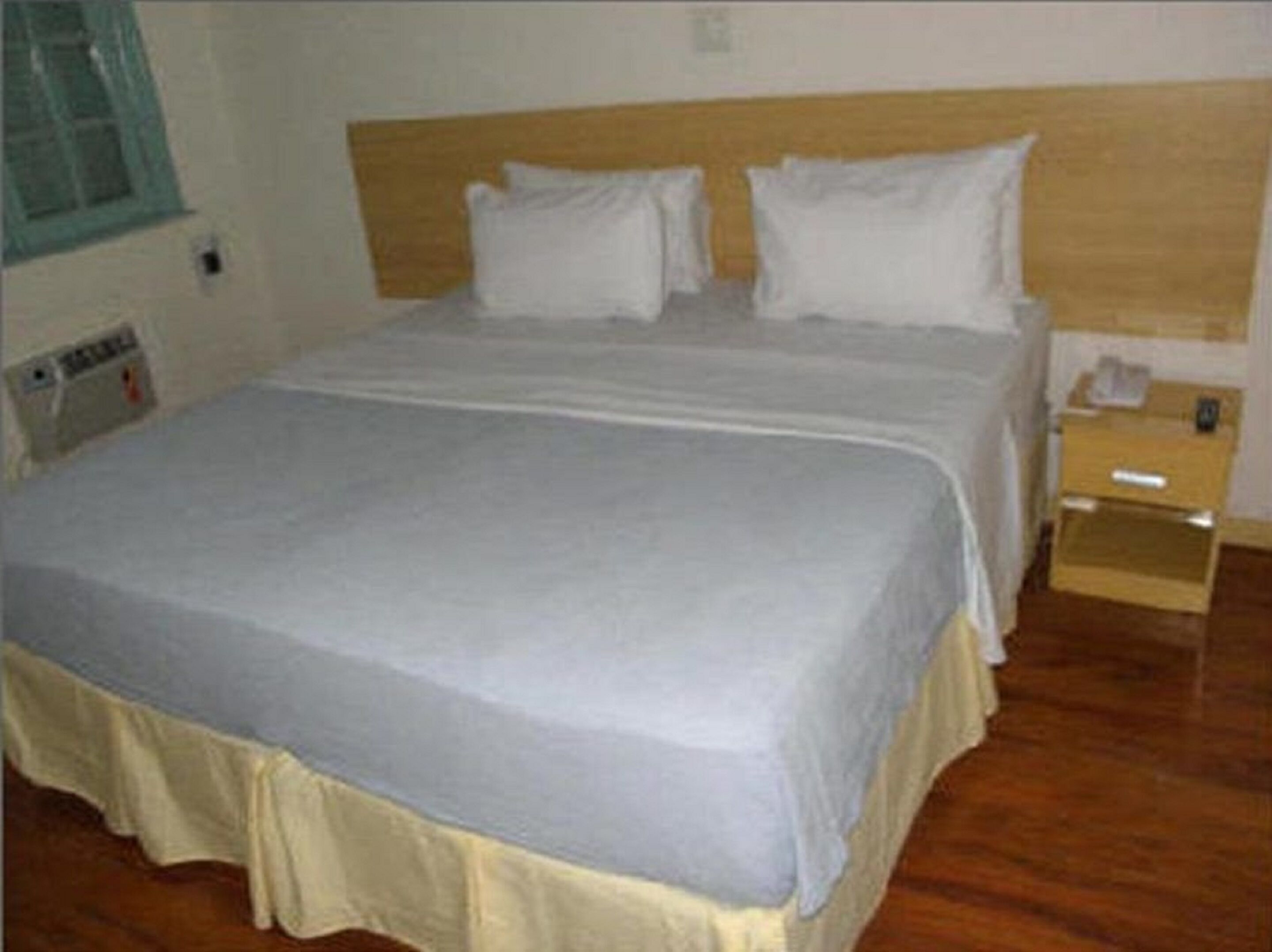 Una cama bien tendida con cabecera de madera, una mesita de noche con lámpara y una ventana con persianas.