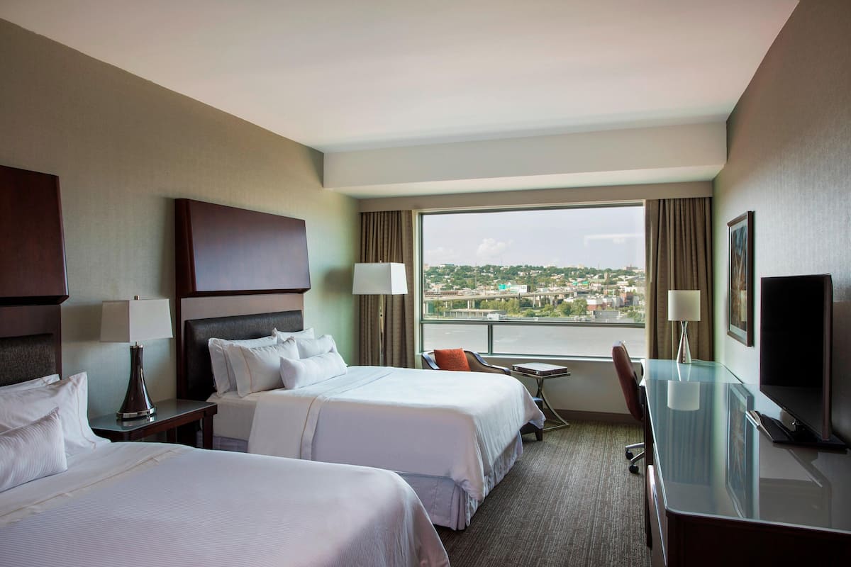 Habitación de hotel con dos camas, un escritorio, un televisor y vistas a un paisaje urbano.