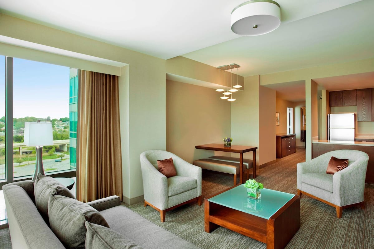 Habitación de hotel con un ventanal grande, un sofá, dos sillones, una mesa de centro y un área de comedor con una mesa y sillas.