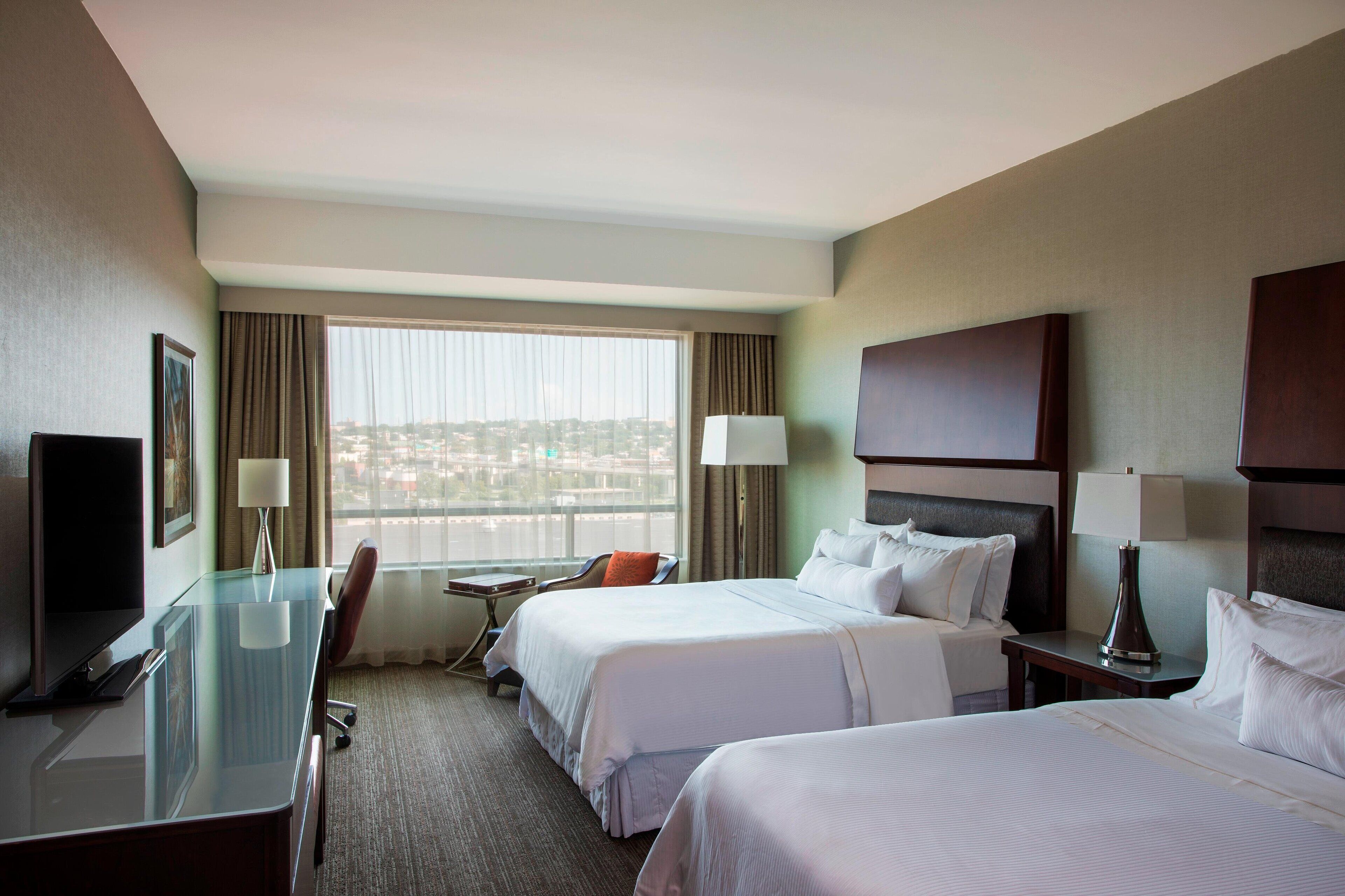 A hotel room with two beds, a desk, a chair, a TV, and a window with a city view.