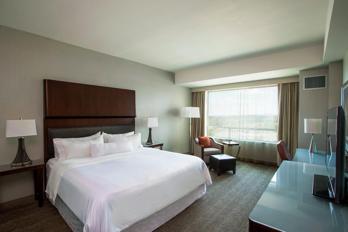 Habitación de hotel con una cama grande, dos mesitas de noche con lámparas, un escritorio con una silla, un televisor y una ventana con cortinas.