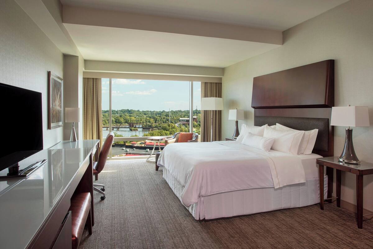 Habitación de hotel con una cama grande, un escritorio con televisor, una silla y vistas a un lago.