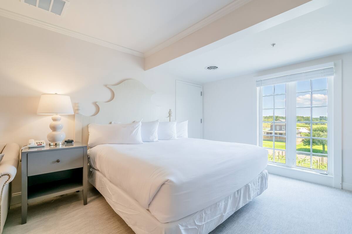 A bedroom with a large bed, a bedside table with a lamp, and a window with a view of the outdoors.