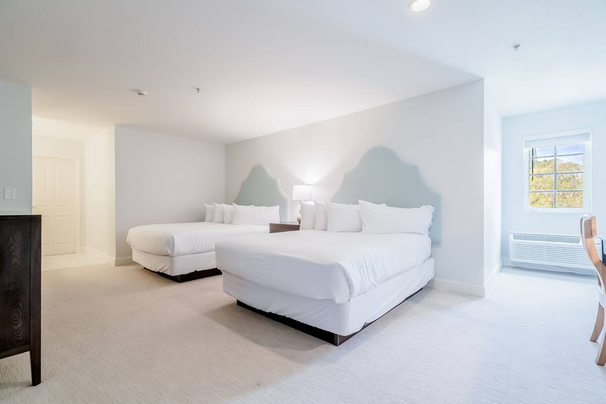 A bedroom with two beds, a nightstand, a lamp, and a window with a view.