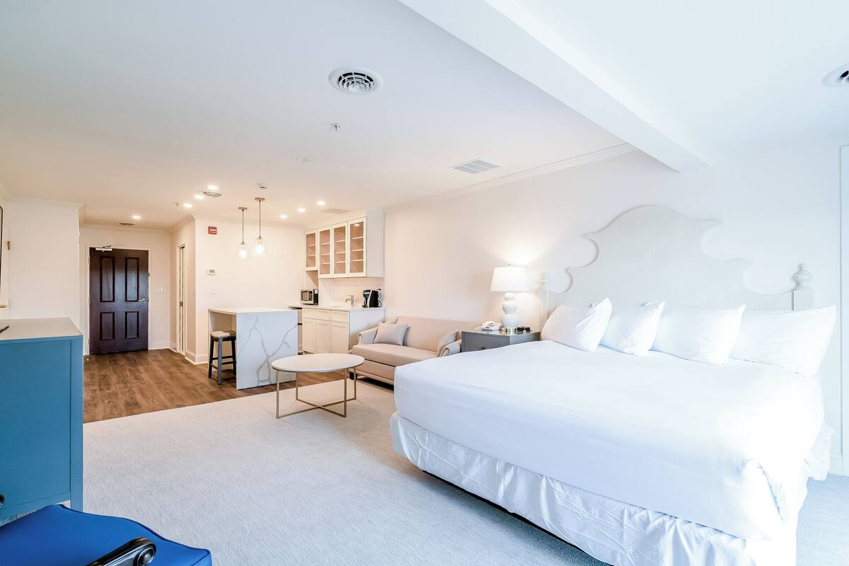 A modern hotel room with a large bed, a desk, a sofa, and a kitchenette.