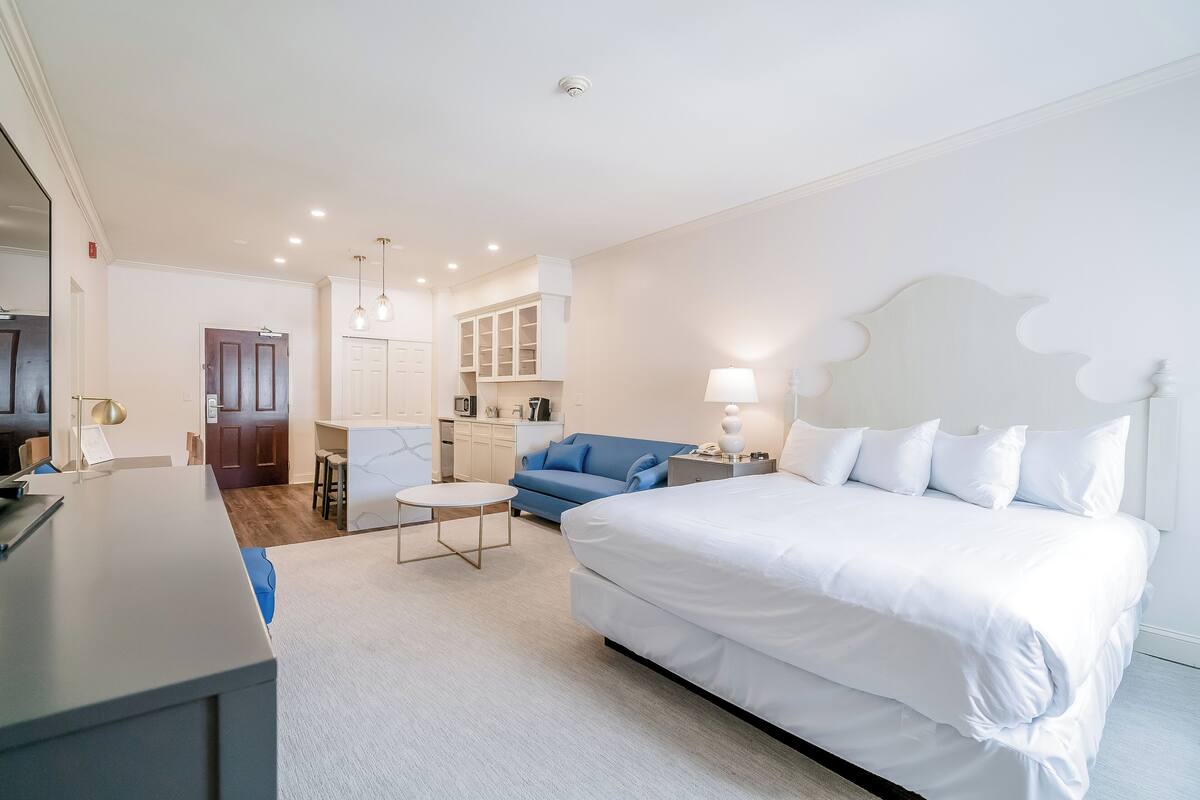 A hotel room with a large bed, a desk, a chair, a small table, a sofa, and a kitchen area.