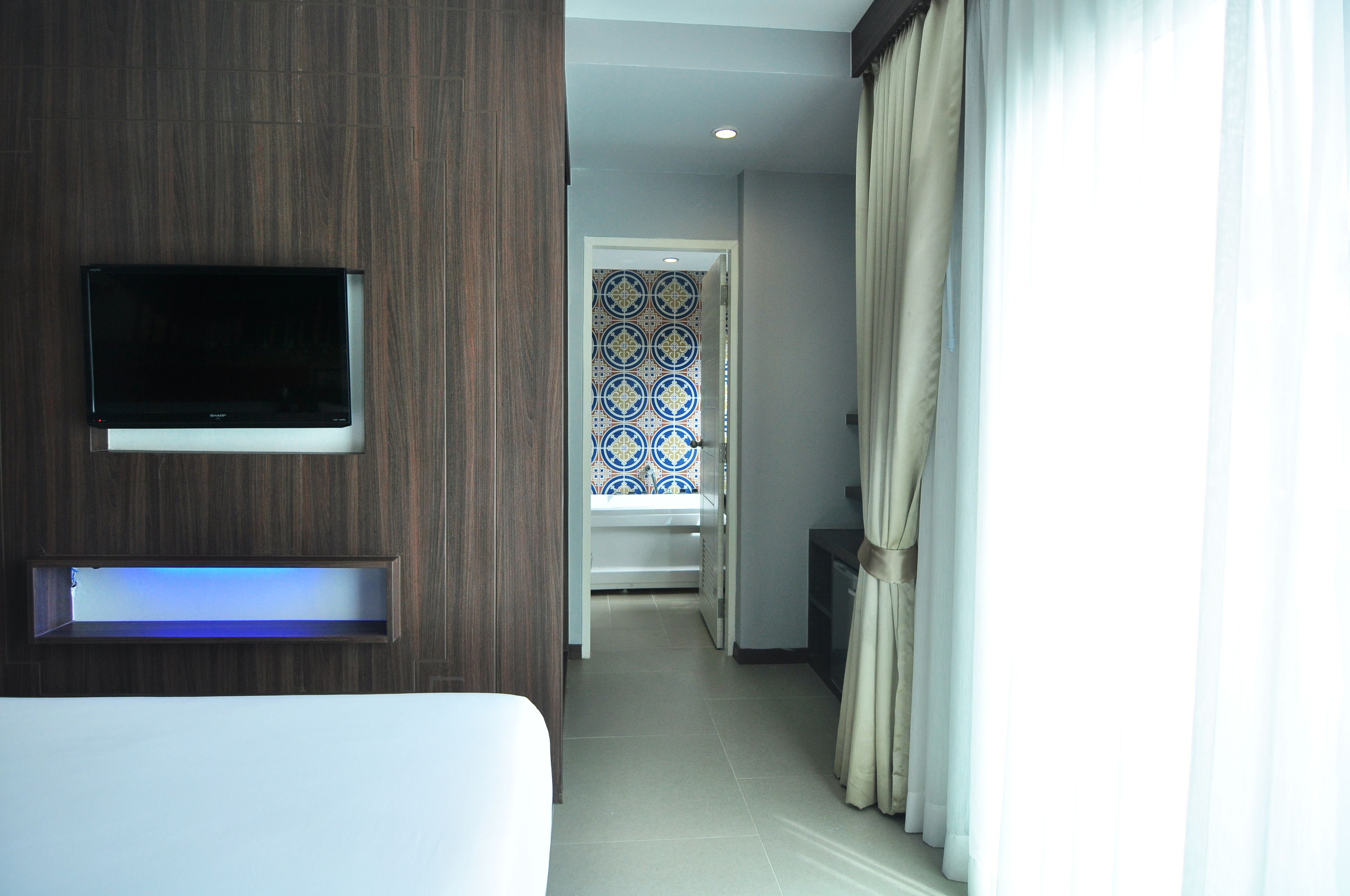 A hotel room with a TV mounted on the wall, a bedside table, and a view of a hallway with a patterned door and curtains.