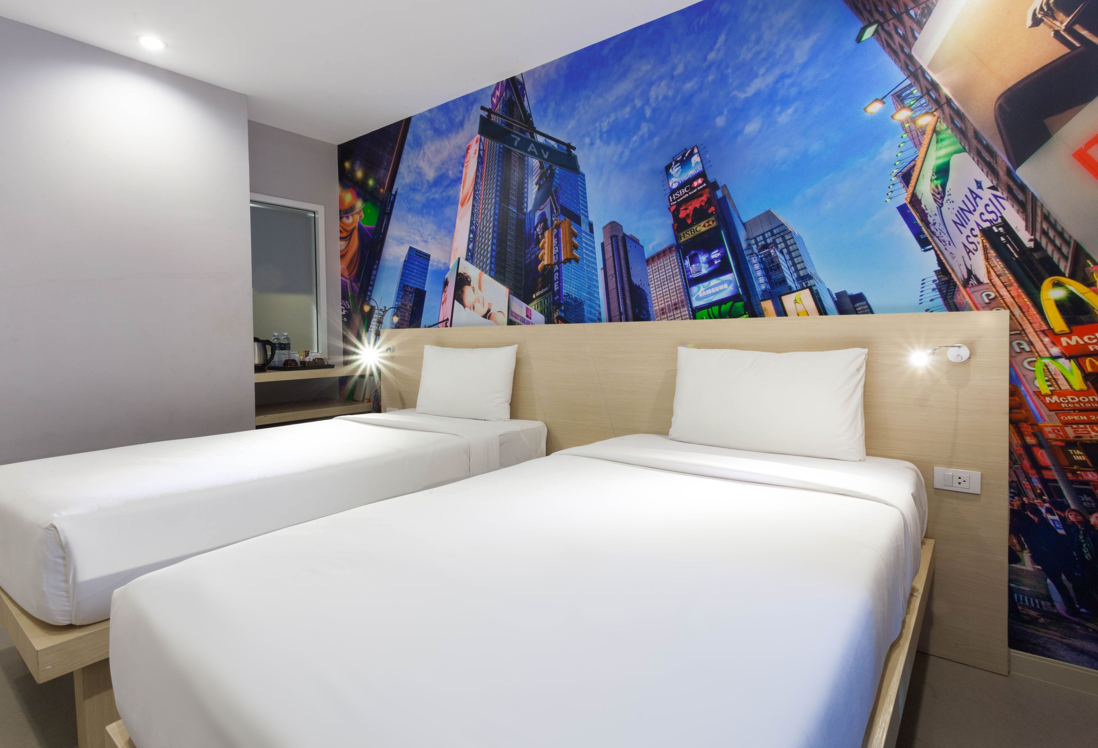 A hotel room with two beds, a large cityscape mural on the wall, and a small table with a kettle.