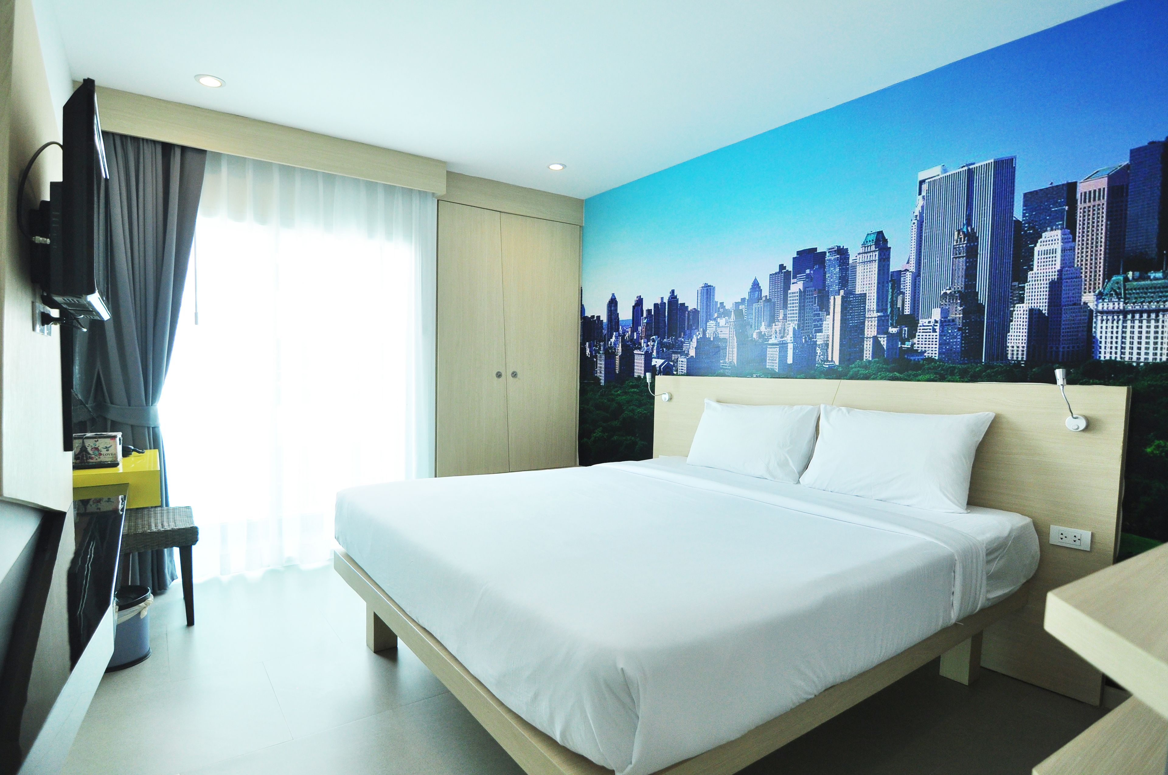 A hotel room with a bed, a television, a desk, and a cityscape mural on the wall.