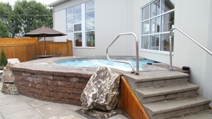 A hot tub with a stone wall and steps leading up to it, a wooden deck, and a wooden fence in the background. - Auberge du Draveur (Maniwaki)
