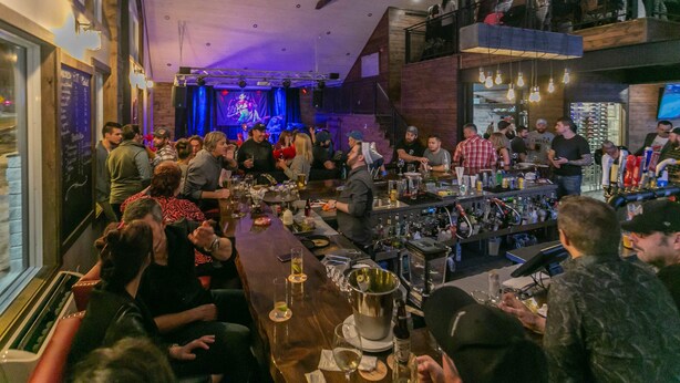 A crowded bar with patrons seated and standing, a bartender at the counter, and a stage with lighting in the background. - Auberge du Draveur (Maniwaki)