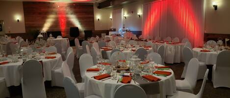 A banquet hall with round tables set for a formal event, white chairs, and a stage with red lighting.
