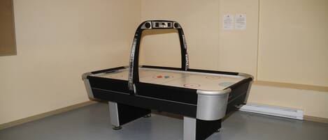 A table hockey game setup in a room with a light-colored floor and walls, featuring a black and silver table with a hockey puck and goals.