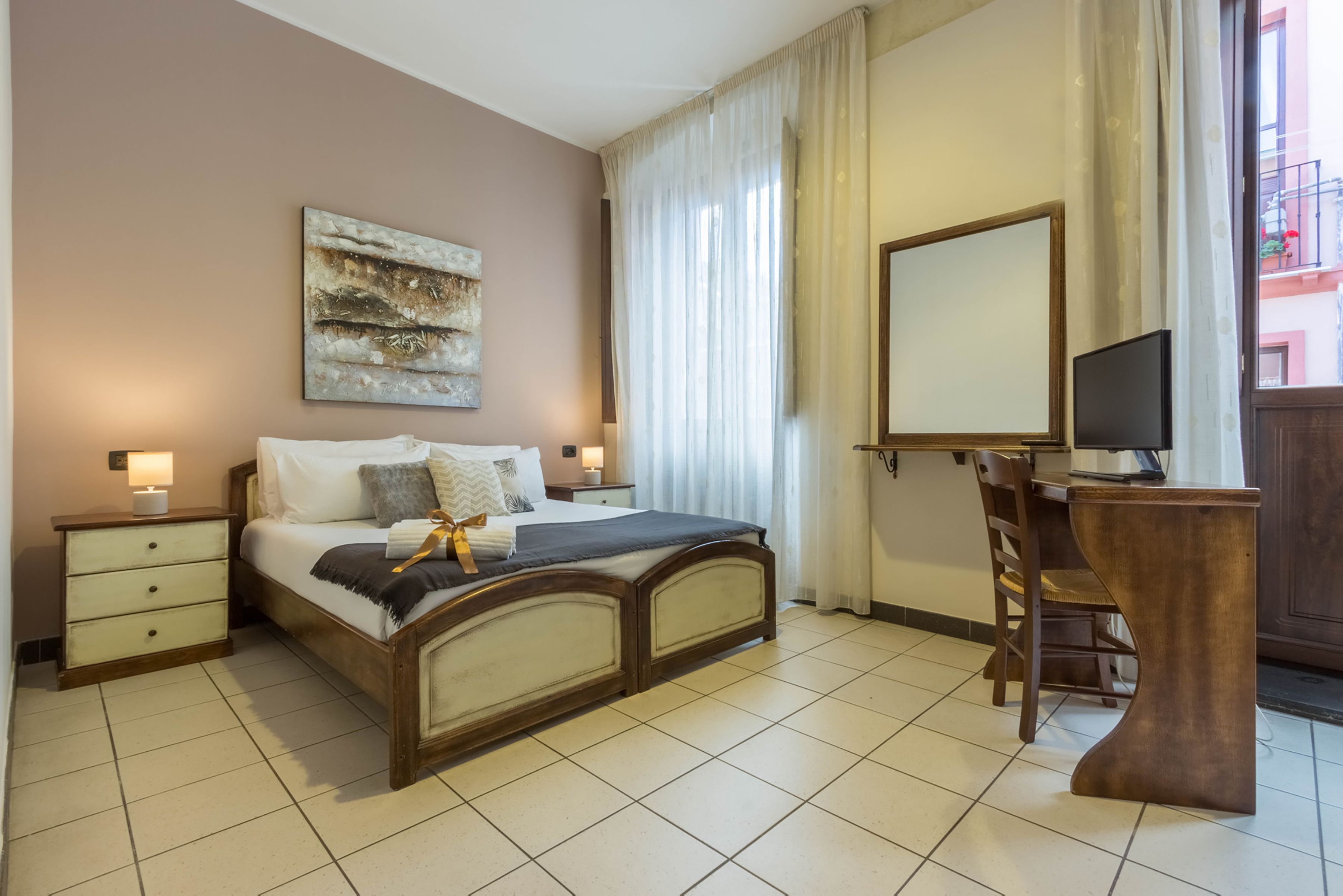 A bedroom with a bed, bedside tables, a desk, a chair, a mirror, and a window with curtains.