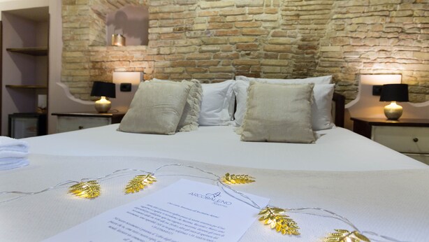 A neatly made bed with white linens, two bedside tables with lamps, a stone wall, and a small shelf. - Arcobaleno Rooms (Cagliari)