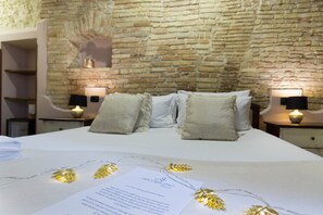 A neatly made bed with white linens, two bedside tables with lamps, a stone wall, and a small shelf. - Arcobaleno Rooms (Cagliari)