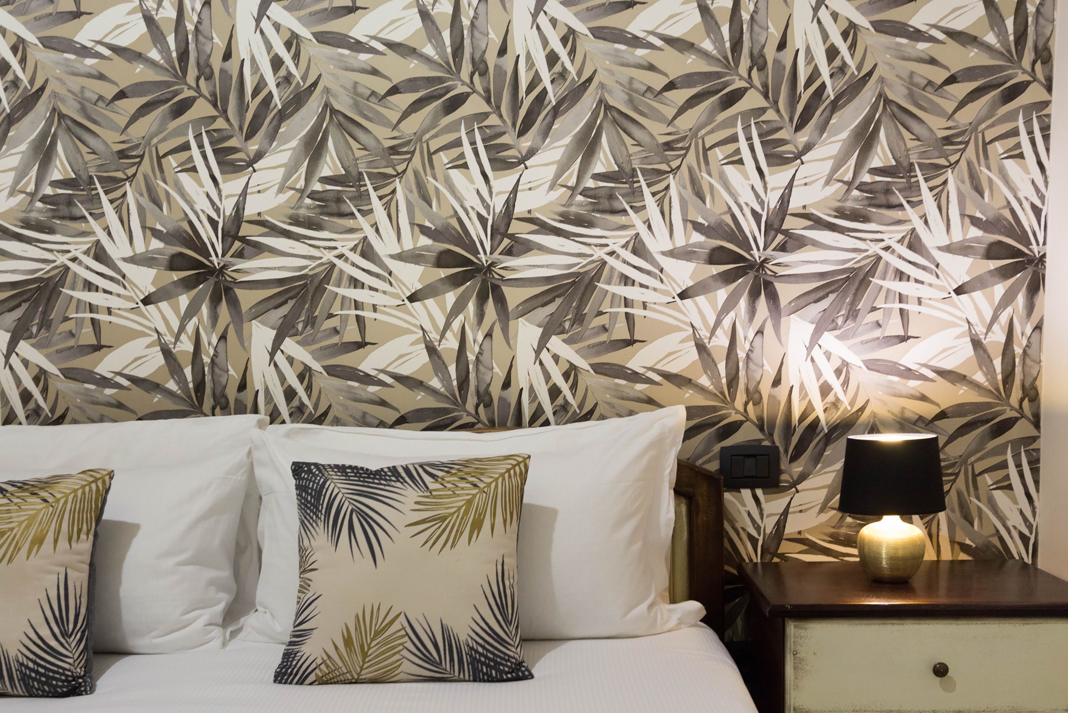 A hotel room with a bed, two pillows, a nightstand with a lamp, and a wall with a leaf pattern.