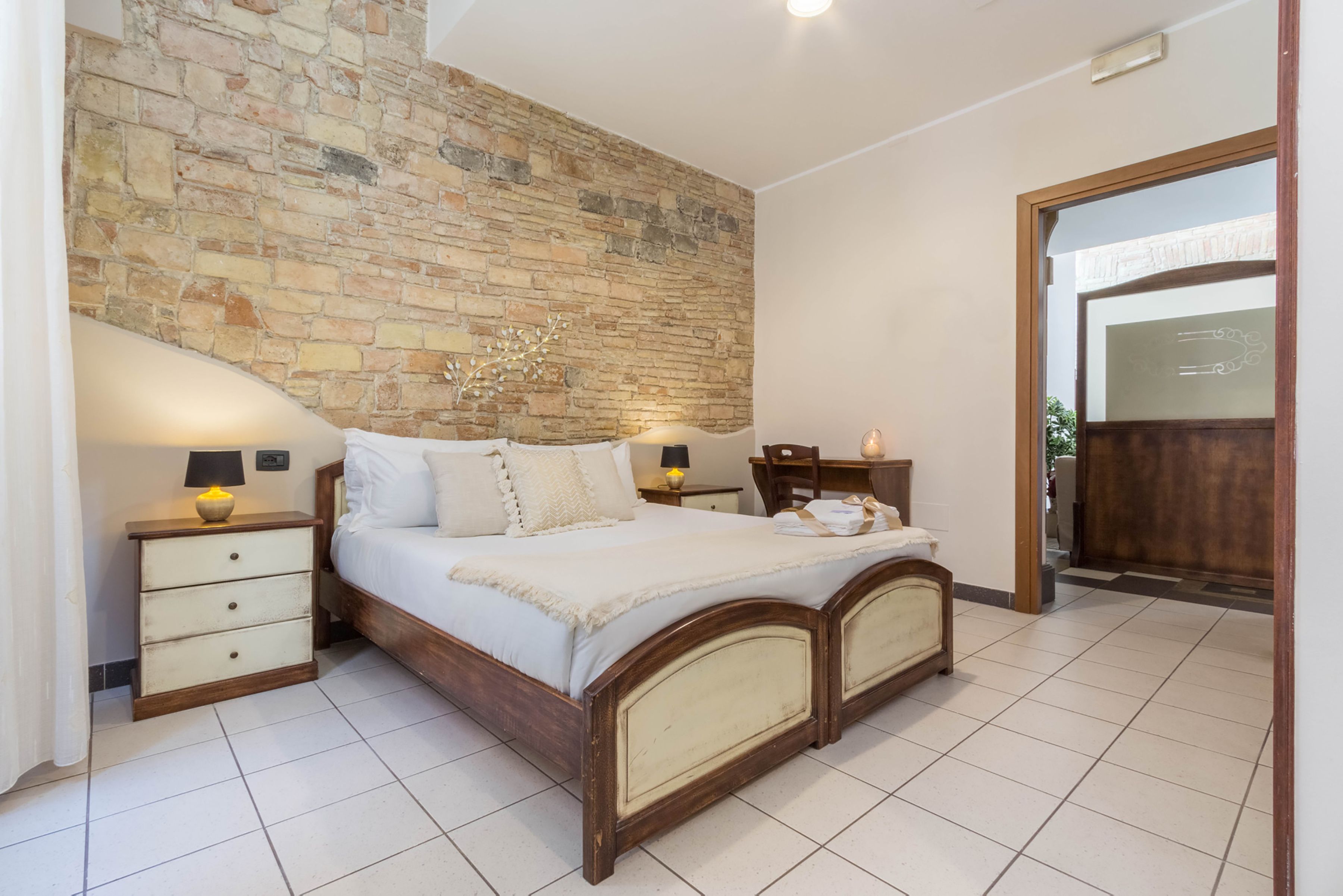 Une chambre avec un mur en pierre, un lit en bois, deux tables de chevet avec des lampes et une porte en bois.