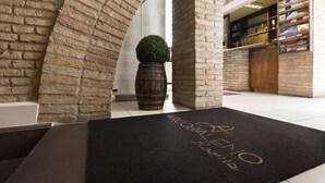 A reception area with a black doormat, brick walls, a wooden barrel, and a small potted plant. - Arcobaleno Rooms (Cagliari)
