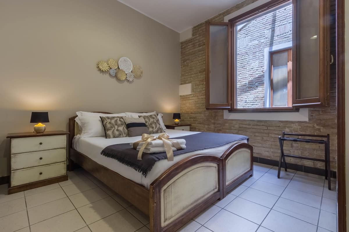 Ein Schlafzimmer mit einem Holzbett, zwei Nachttischen mit Lampen, einem Fenster mit Backsteinwand und Wanddekoration.