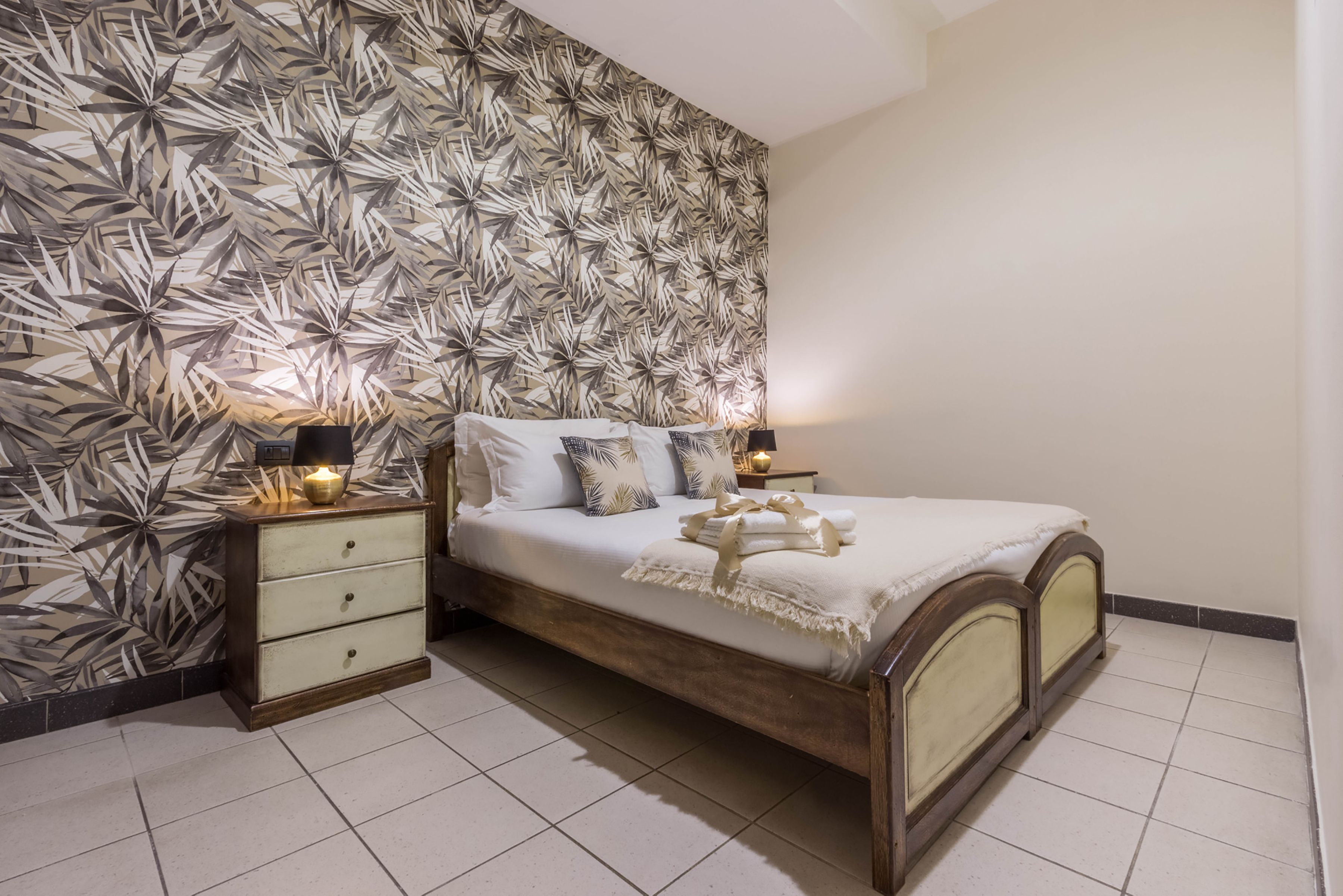 A bedroom with a patterned wallpaper, a wooden bed frame, two bedside tables with lamps, and a nightstand with drawers.