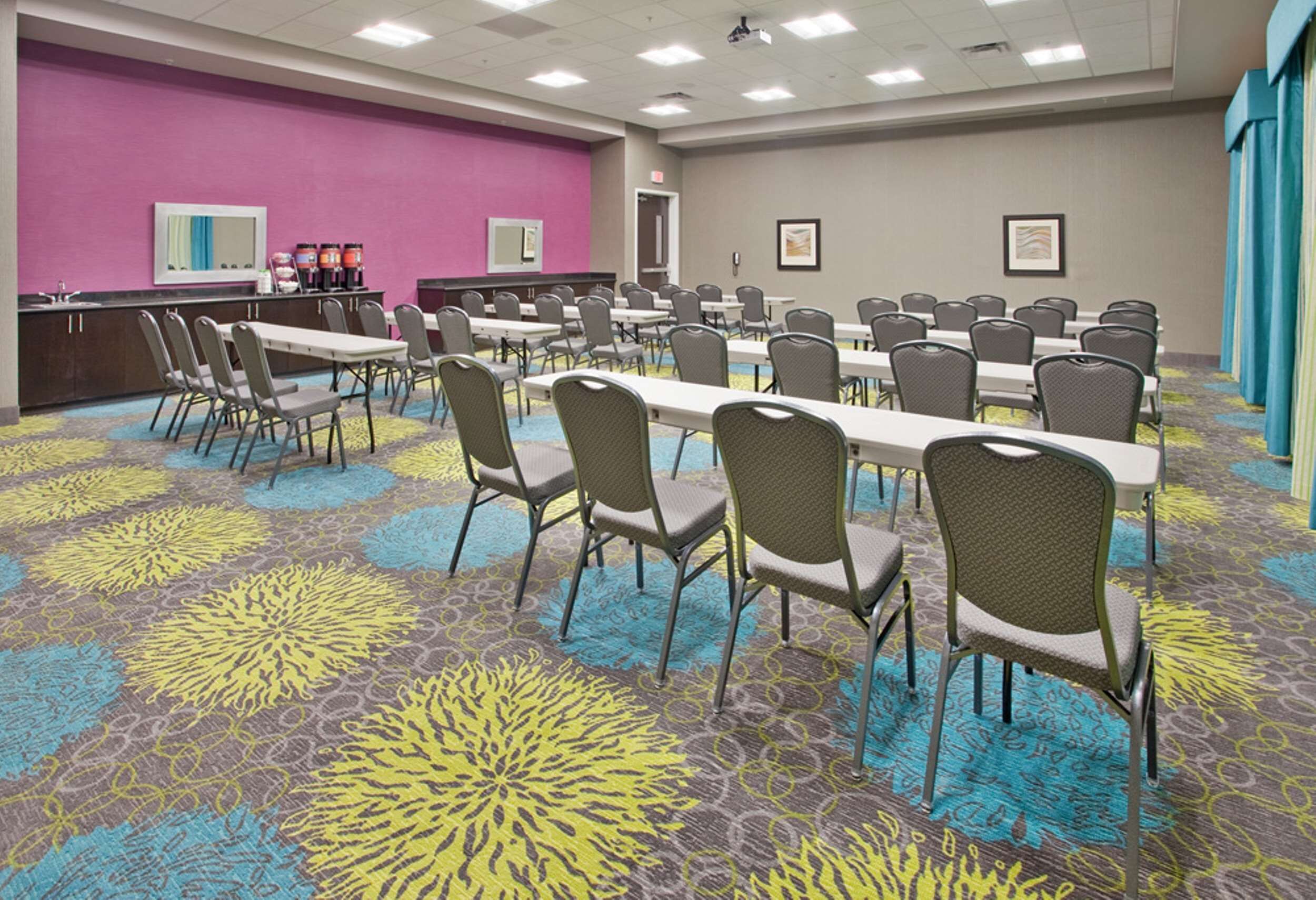 A conference room with multiple tables and chairs, a coffee station, and a colorful carpet.