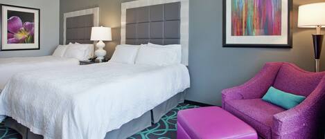 A hotel room with two beds, a purple armchair, a colorful abstract painting, and a framed floral artwork.