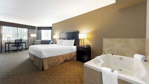 A hotel room with a large bed, a desk, a chair, a desk lamp, a nightstand with a lamp, and a bathtub. - Best Western Pembroke Inn & Conference Centre (Pembroke)
