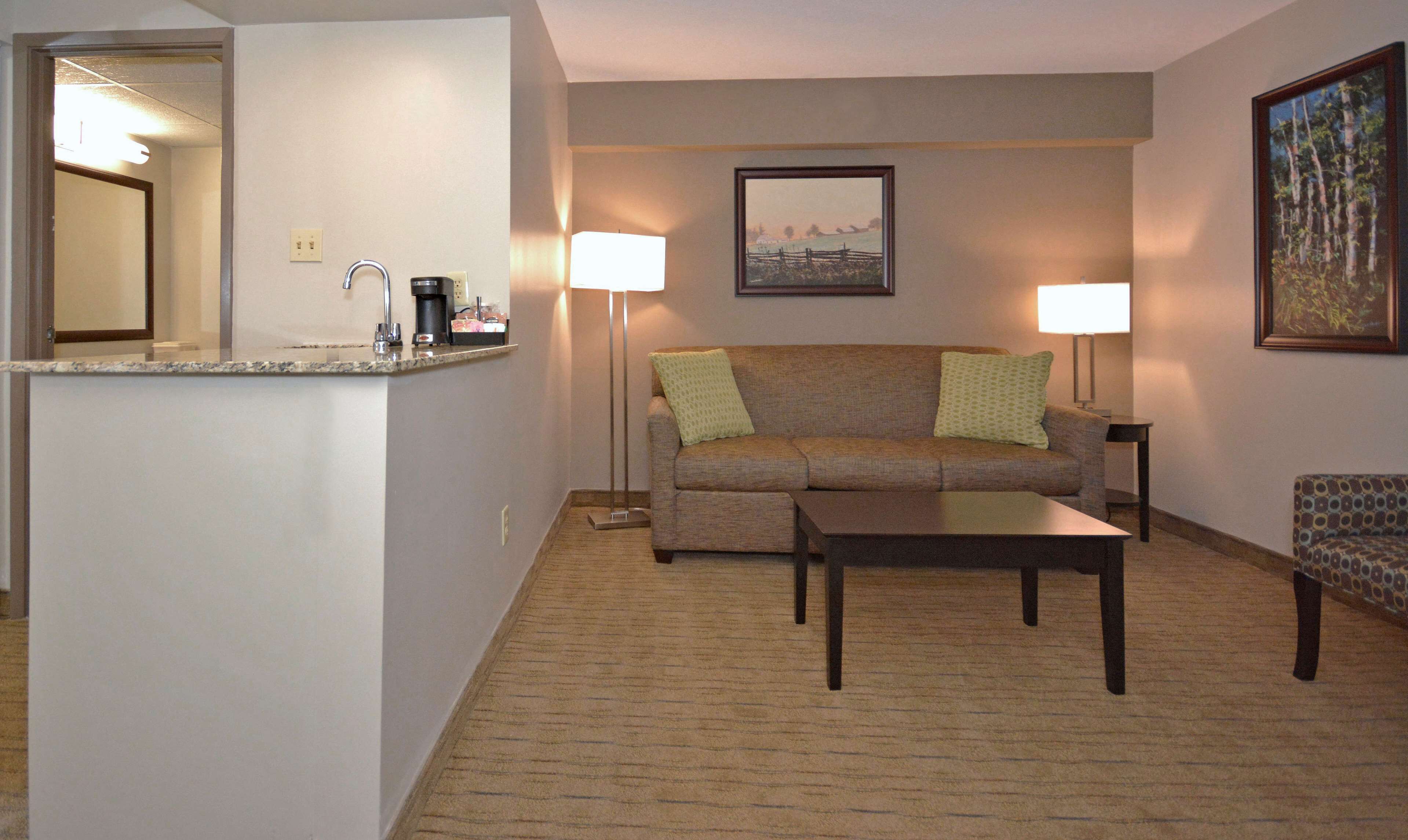 A living room with a sofa, a coffee table, a patterned armchair, and a small side table. There is a kitchen area with a countertop and a coffee machine. Two floor lamps provide lighting.