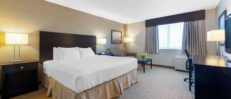 A hotel room with a large bed, bedside tables, a desk, a chair, a TV, and a window with curtains.
