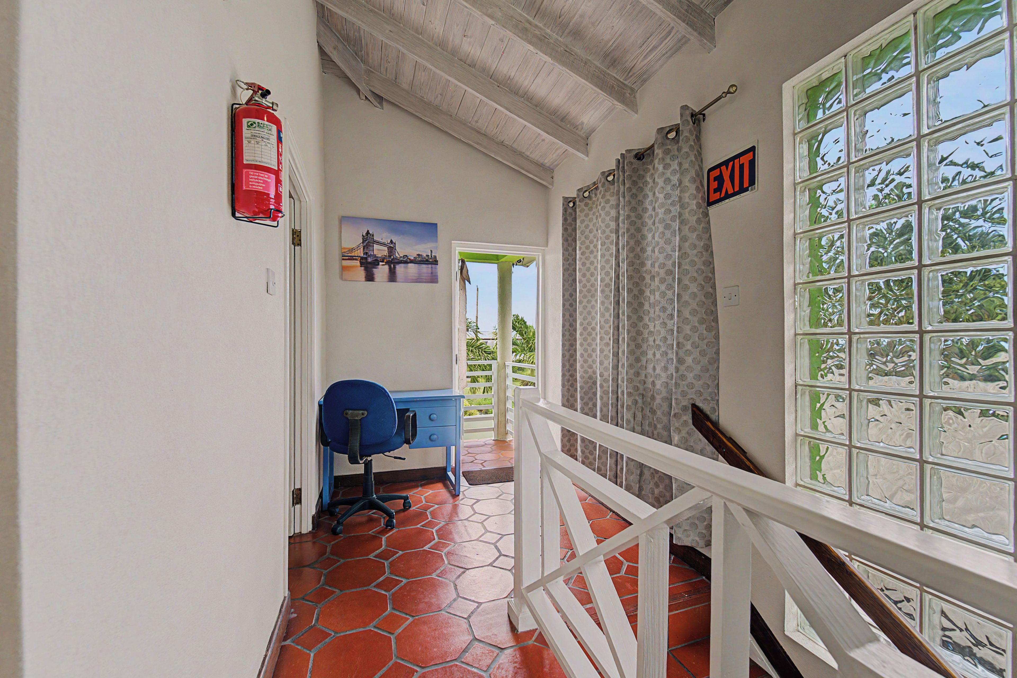 A hallway with a blue office chair, a fire extinguisher, a painting of a bridge, and an exit sign.