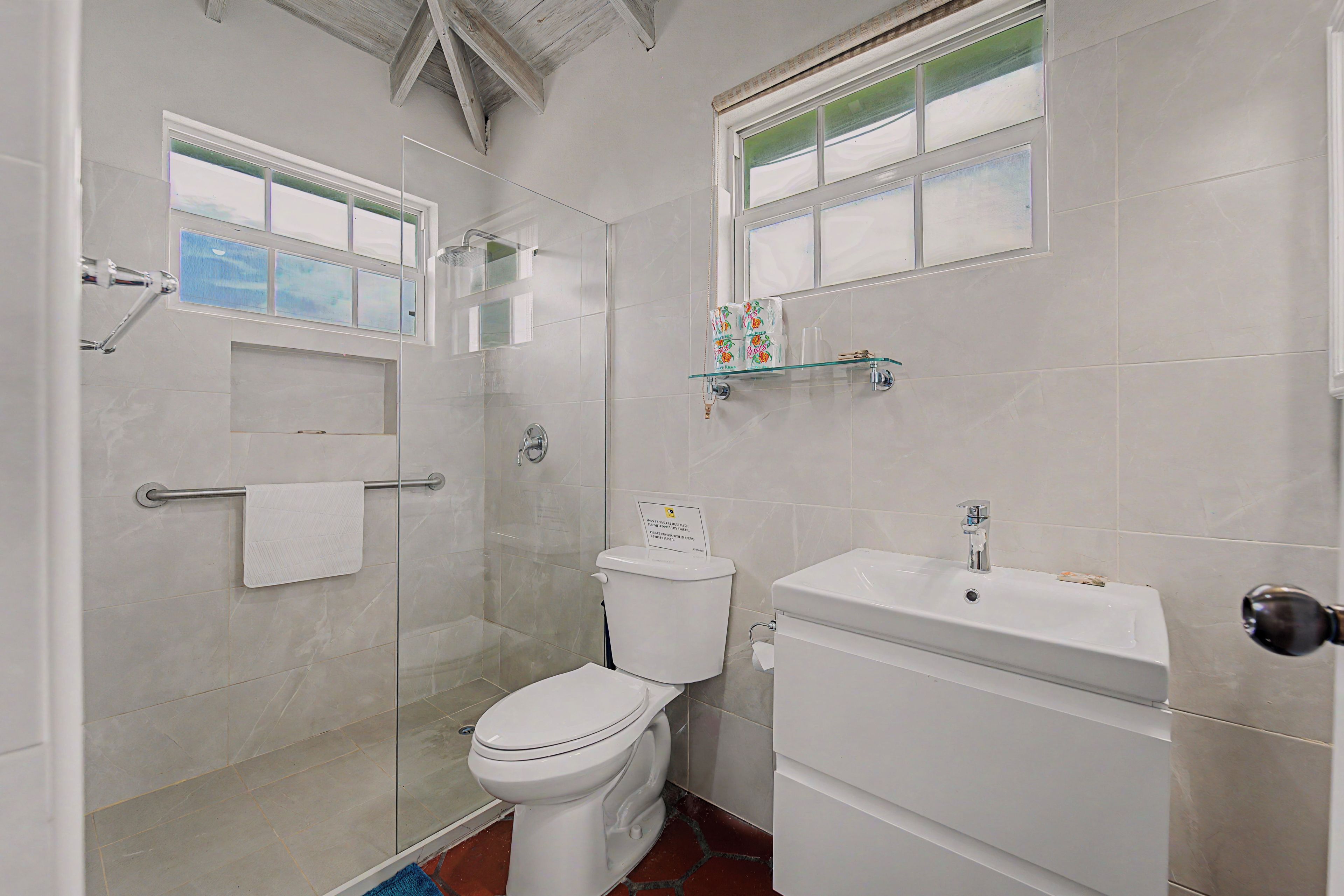 Un baño con ducha de vidrio, inodoro, lavamanos y ventana sobre el lavamanos.