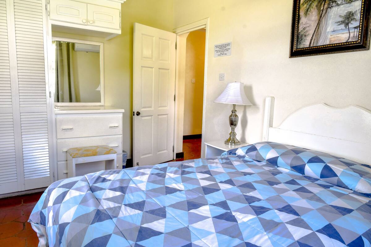 Une chambre à coucher comprenant un lit, une table de chevet avec une lampe, une commode et une porte donnant sur une autre pièce.