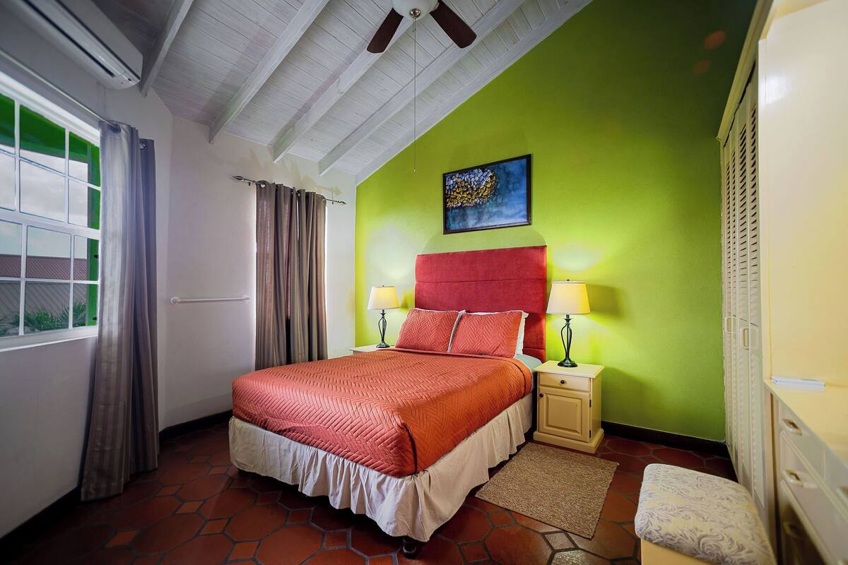 Une chambre d’hôtel avec un lit, des tables de chevet, une fenêtre avec des rideaux, un ventilateur de plafond et un mur vert.