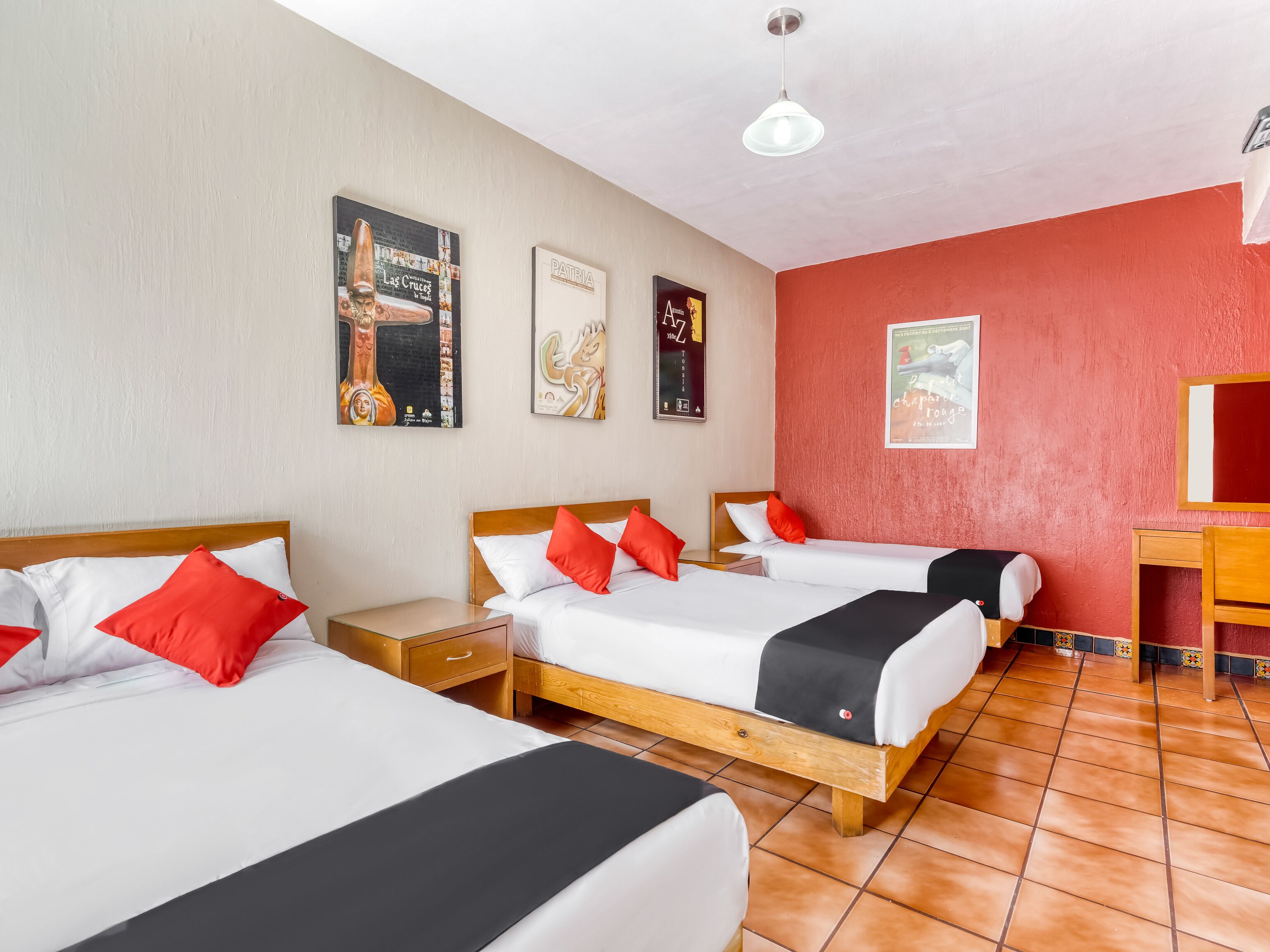 Hotelkamer met drie enkele bedden, elk voorzien van witte lakens en rode kussens. Er zijn houten nachtkastjes, een klein bureau met een stoel, en een rode accentmuur.