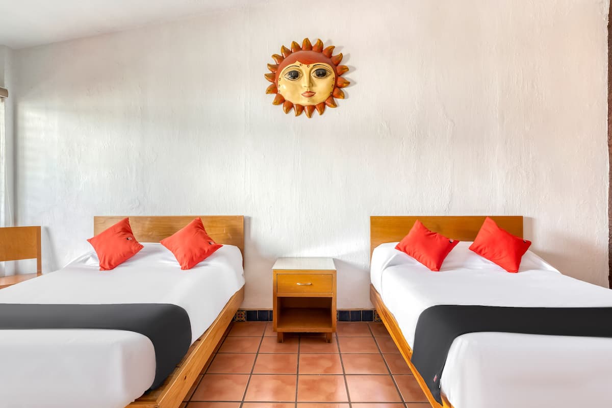 Habitación de hotel con dos camas, cada una con cabecero de madera, ropa de cama blanca y almohadas rojas. Entre las camas hay una mesita de noche de madera, y arte decorativo con forma de sol en la pared sobre el cabecero.