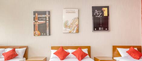Habitación de hotel con dos camas individuales y una cama doble, cada una con ropa blanca y almohadas rojas. Hay mesitas de noche de madera con cajones, y el suelo está cubierto de baldosas. Tres carteles decoran la pared sobre las camas.