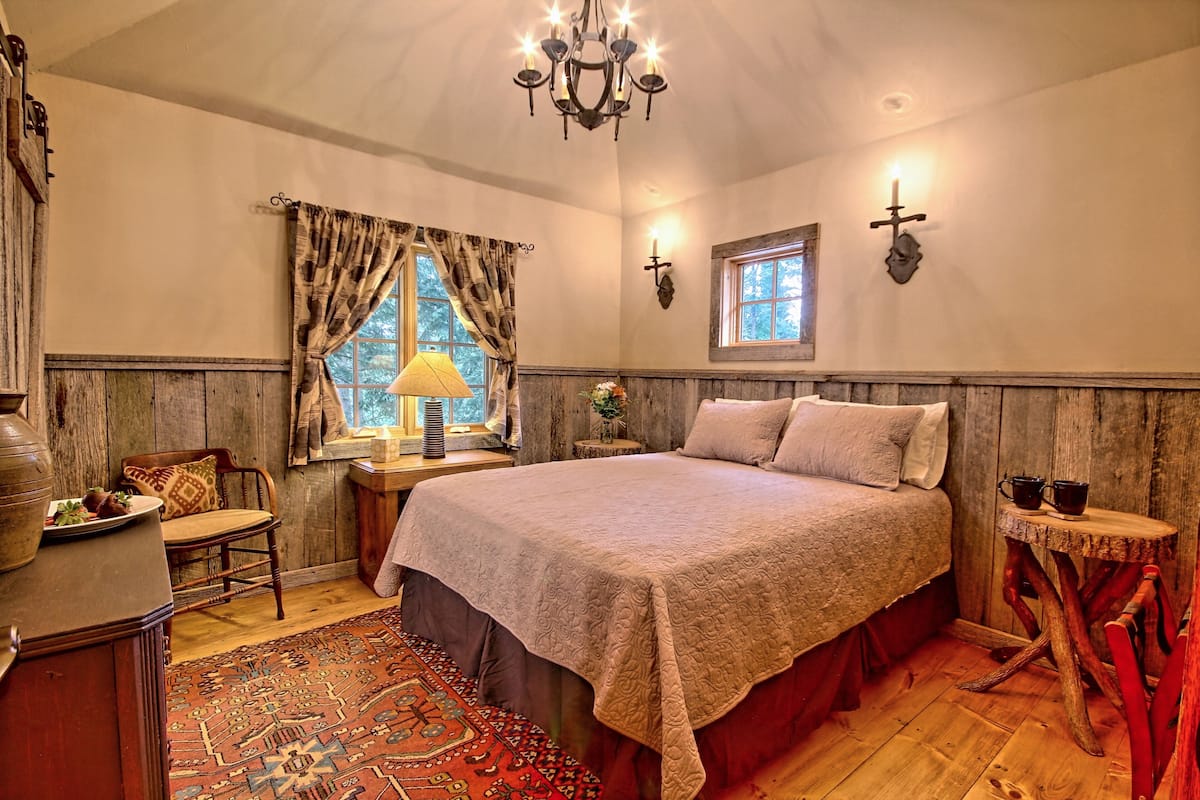 A bedroom with a large bed, wooden walls, a chandelier, a window with curtains, and a small table with a lamp.